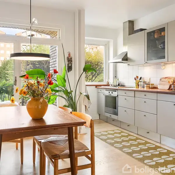 Et køkken med grå skabe og en komfur, bord med gule vase med blomster, bænke med puder, og store vinduer viser en balkon med planter. Farvede lysguirlander hænger ved døren.