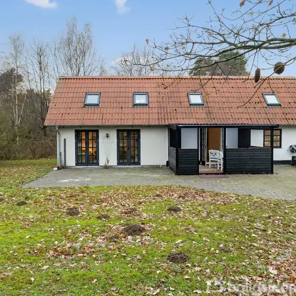 Et hvidt hus med rødt tegltag står stille i en frodig have omgivet af træer. Huset har flere vinduer og en mørk veranda med et bord og stole.