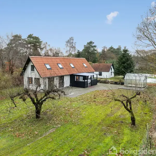 Hus med rødt tegltag og tagvinduer står på en græsplæne med nøgne træer. Baggrund viser en have og et drivhus i en skovfyldt område under blå himmel.