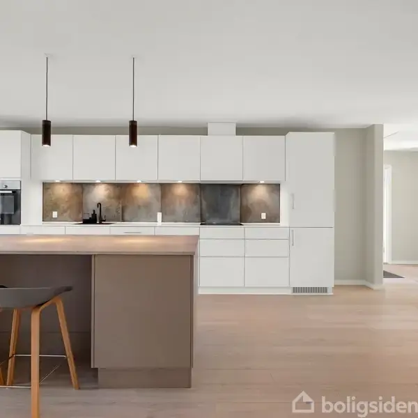 Køkken med hvidt skab, brun køkkenø med barstole og loftlamper i moderne stil. Blomster i vase på bordet, trægulv og neutral vægfarve i baggrunden.