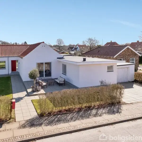 Et hvidt enfamiliehus med rødt tag og garage står på en flisebelagt gårdsplads. Til venstre er en velplejet græsplæne, omkranset af buske. Solrig boligkvarter med flere huse.