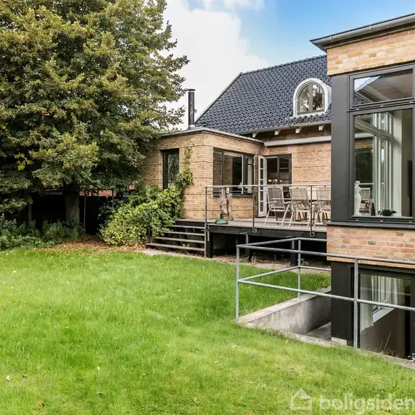 En terrasse af træ med borde og stole står på en forhøjet del af en murstensbygning, omgivet af en grøn plæne og buske under en blå himmel.