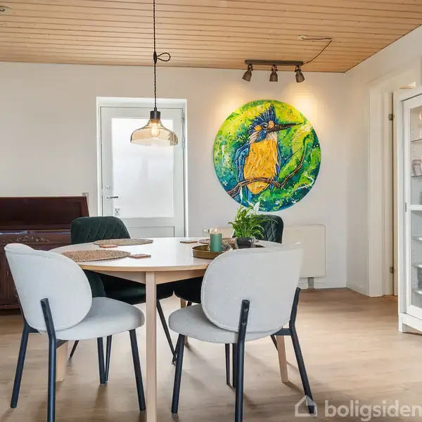 Rundt bord med fire stole står i en spisestue med lyse trægulve. Vægdekorationer inkluderer en stor, farverig ugle og et abstrakt maleri. Glasskab viser porcelæn.