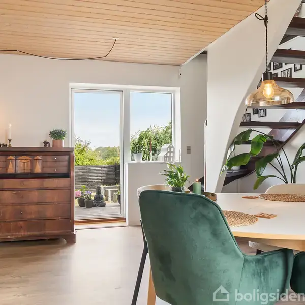 Et spisebord med fire grønne stole står i et lyst rum. Ved siden af er en trappe, og en stor plante pynter ved væggen. En glasskab og et antikt skab ses til venstre, mens en glasdør leder ud til en terrasse med udsigt over en have.
