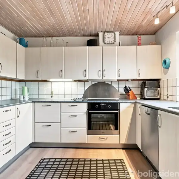Et moderne køkken med lyse træskabe. En indbygget ovn og komfur er placeret centralt. På bordet står en toaster og et skærebræt. Loftet har træpaneler og indbygget belysning.