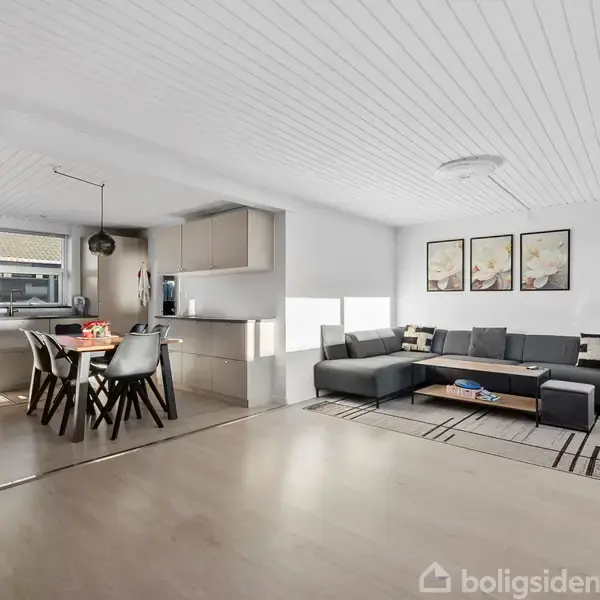 Et moderne køkken-alrum med et spisebord og seks stole til venstre, og en grå hjørnesofa til højre. Lyse vægge og gulve skaber en rummelig atmosfære.