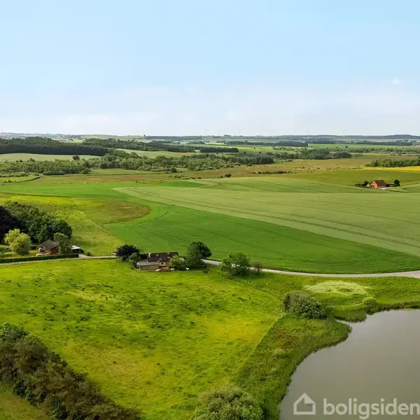 Landskab med marker og en lille dam. En vej løber igennem området, omgivet af grønne træer og åbne vidder, med enkelte bygninger spredt i det fjerne.