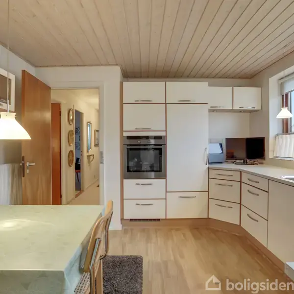 Køkken med hvide skabe har ovn og kogeplade. Spisebord med to stole står under en lampe. Vinduet ved siden af vasken giver udsyn til et træ udenfor.