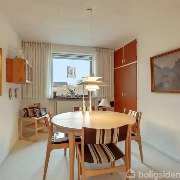 Rundt spisebord omgivet af fire stole med stribede hynder, placeret i spisestue med malerier på væggene og et vindue med udsigt. Lampe hænger over bordet.