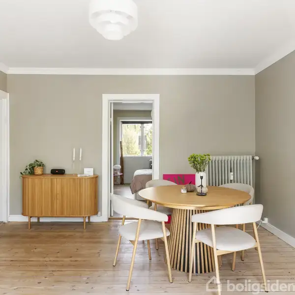 En rund træbord med hvide stole omkring står i en stue med lysegrå vægge. Et skab med planter og bøger står i baggrunden ved siden af en radiator.