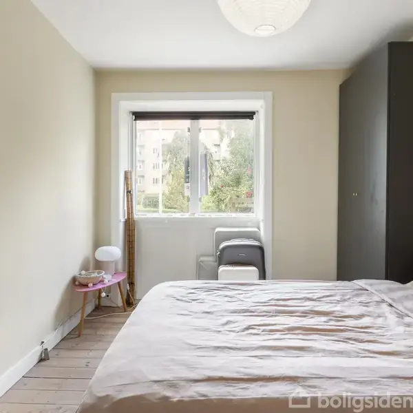 Et soveværelse med en dobbeltseng dækket af lyserødt sengetøj. Der står et rundt bord med en lampe ved siden af sengen. Vindue med udsigt til træer.