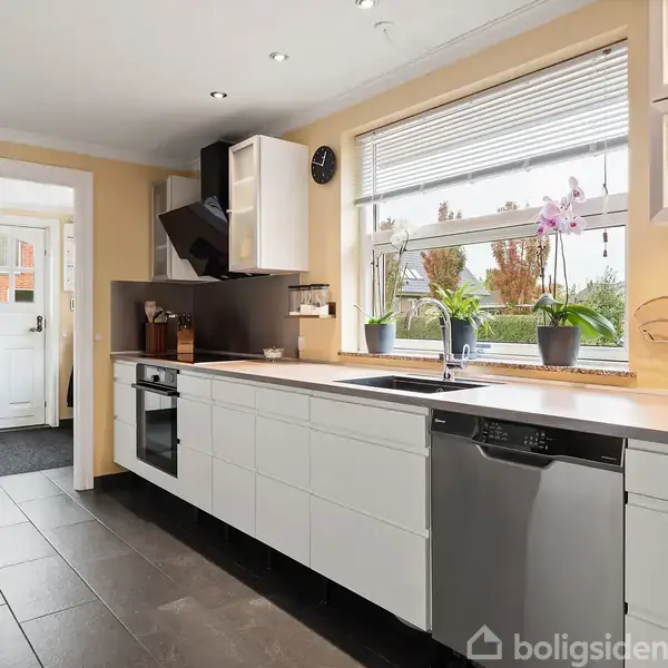 Et moderne køkken med hvide skabe, en indbygget ovn og opvaskemaskine. En stor vindueskarm med planter og orkideer giver udsigt til en have. Døren fører til entréen.