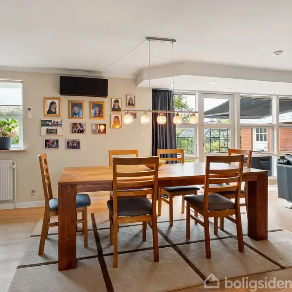 Spisebord med seks stole står under en række hængende lamper i en lys stue. Der er fotografier på væggen og store vinduer med udsigt til en terrasse.