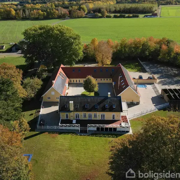 Central gård bygning omkranser græsplæne, beliggende i frodigt, landskab. Omgivet af træer, med åbne marker i baggrunden og flere strukturer i nærheden.