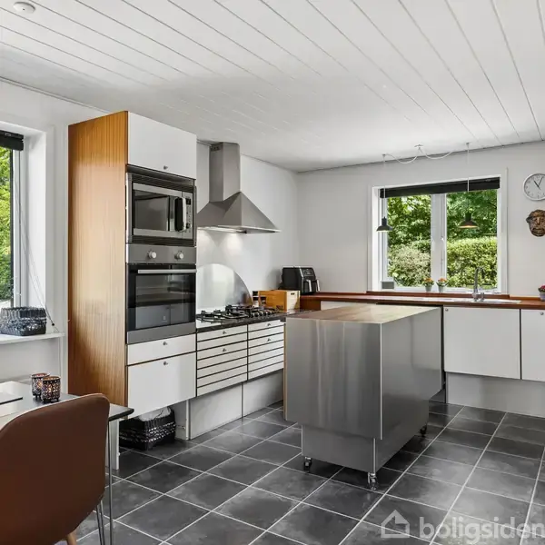 Køkken med en køkkenø i stål, ovn, komfur og emhætte; to vinduer slipper naturligt lys ind. Gråt flisegulv og hvidmalet loft. Spisebord med brune stole til venstre.