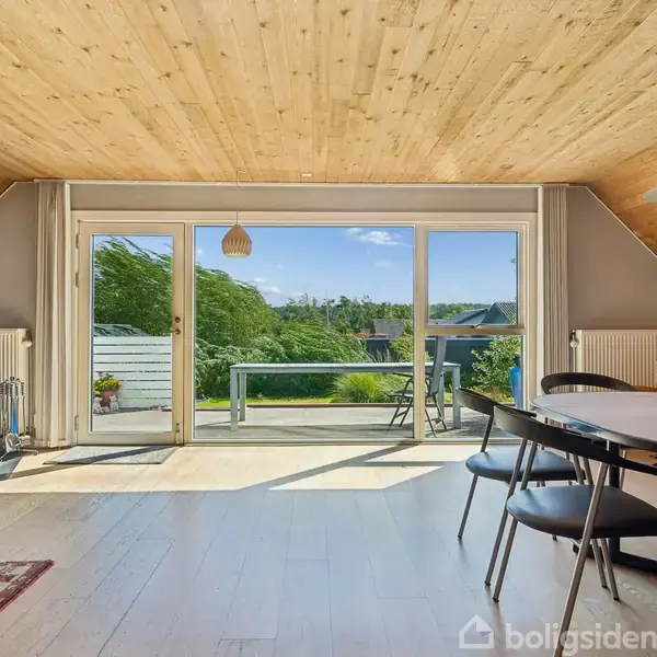 Brændeovn ved siden af glasdøre, udsigt til terrasse og grønne omgivelser. Spisebord med stole på lyse trægulve i rum med skrå loftplader. Sollys skinner ind gennem dørene.