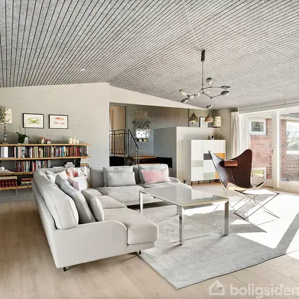 En lys stue med en grå sofa og sofabord i centrum. Hylder med bøger bag sofaen. Store vinduer giver udsigt til en have, og der hænger en moderne lampe i loftet.