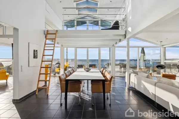 En spisebord med brune læderstole står i et lyse og moderne rum med store glasvægge, der giver udsigt til havet. En stige fører op til en åben hems.