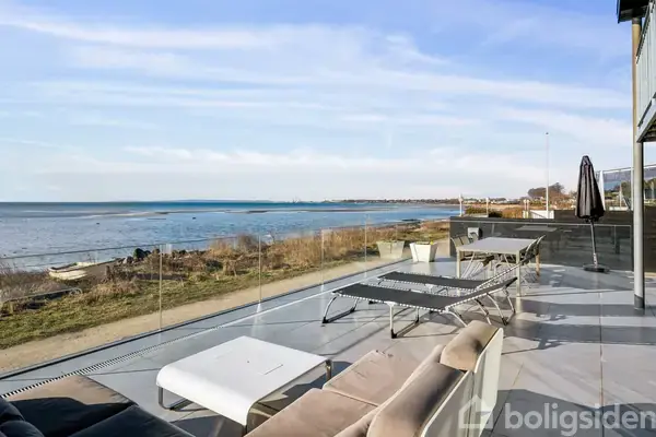 Terrasse med sofa og borde står stille. Udsigt til en fredelig kystlinje og strand med græs og hav. Klar blå himmel giver en solrig atmosfære.