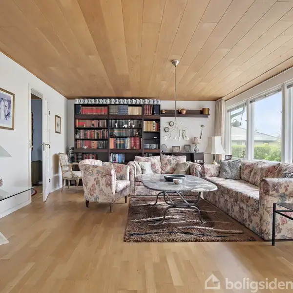 En stue med blomstrede sofaer og stole er arrangeret rundt om et sofabord. En stor reol fyldt med bøger står i baggrunden. Lyse vægge og store vinduer giver lys.
