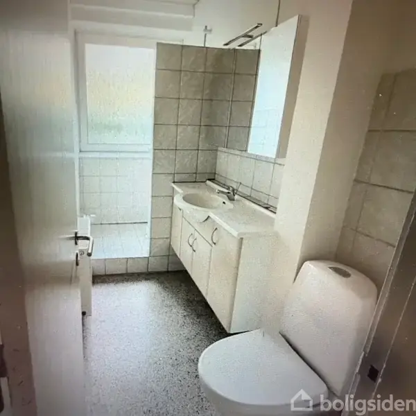 Badeværelse med vask på et hvidt skab, toilet til højre, flisebelagte vægge og gulv, samt en brusekabine med vindue i baggrunden.