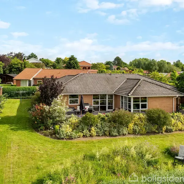 En enkeltstående murstensvilla med sort tag står midt i en frodig have, omgivet af buske og træer. Græsplænen er velholdt, og der er en hængekøje i nærheden.