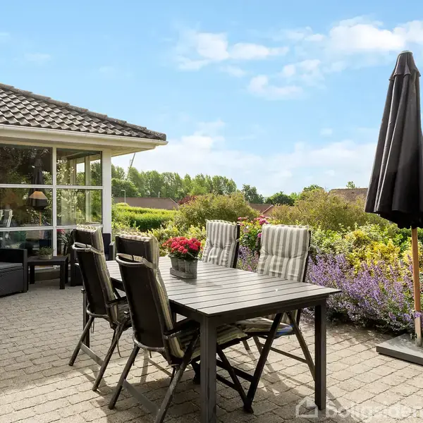 Terrassemøbler står på flisebelagt terrasse ved huset, med bord og stole omkring. En sort parasol beskytter mod solen, omgivet af blomsterbede og grønne buske i tæt bebyggelse.