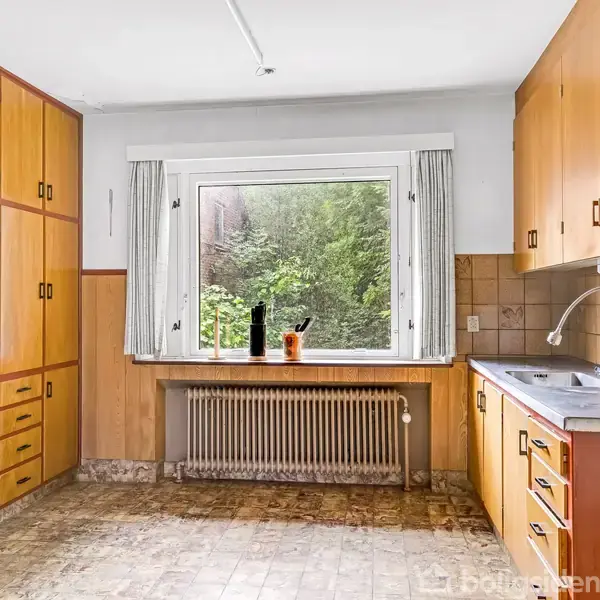 Et køkken med træskabe står tomt. En stor vindue åbner op til en udsigt af grønt løv. En radiator er placeret under vinduet. Installationen har en enkelt vask.