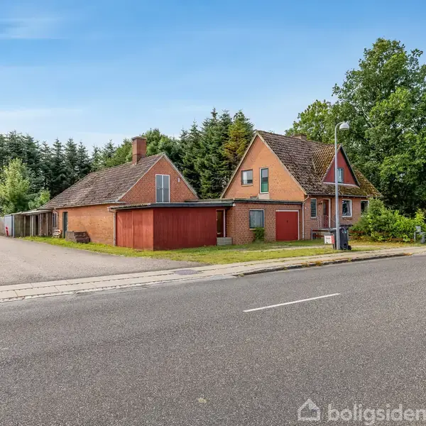 Et rødt murstenshus med saddeltag står langs en vej. Der er træer bagved, og en asfaltvej i forgrunden. Tre redskabsskure er ved siden af huset.