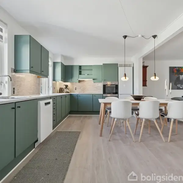 Køkken med grønne skabe strækker sig langs to vægge, med hvidt bordplade og stålvask. Spisebord med seks hvide stole står under tre hængelamper i et lyst rum med parketgulv.