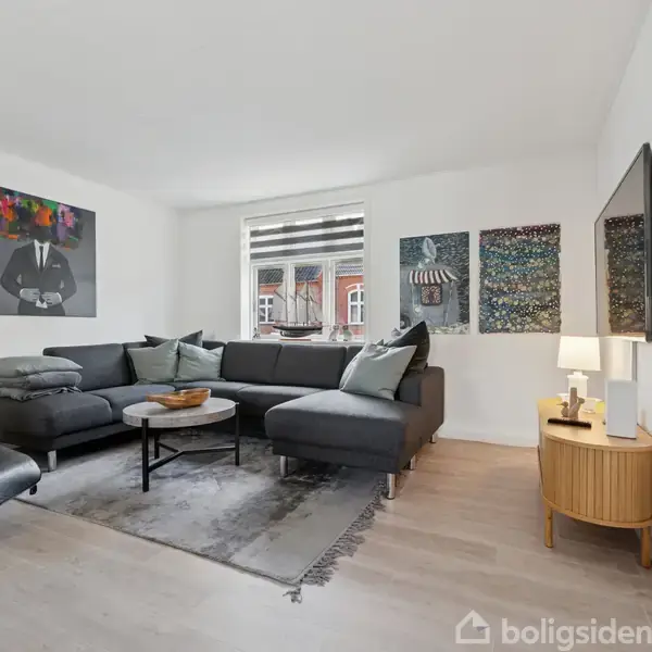 Stor grå hjørnesofa står i stue med lyse vægge og trælaminatgulv. Ved siden af hænger moderne kunstværker. Et lille rundt sofabord står foran sofaen, og fjernsyn hænger på væggen.