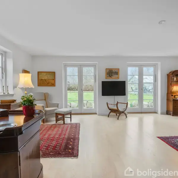 Stue med møbler som lænestole og skrivebord, lyse trægulve, to gulvtæpper, vægmonteret tv, og to sæt franske døre med udsigt til en have. Malerier og lamper dekorere rummet.