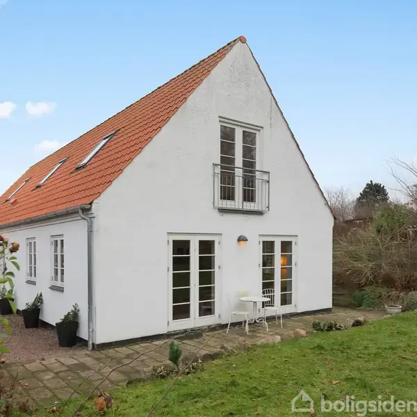 Hvidt hus med rødt tag står omgivet af en have med græsplæne. Husets facade har franske døre og en lille balkon. Himlen er blå med lette skyer.