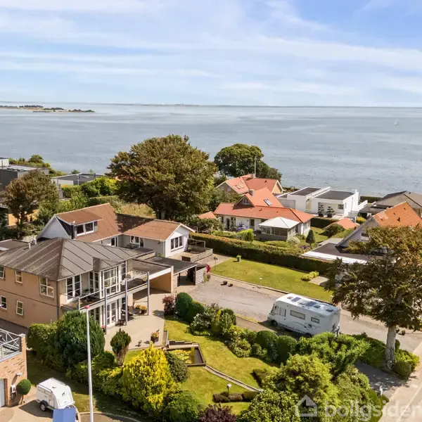 Huse ved kysten har velplejede haver, mens en campingvogn holder parkeret på en vej i nærheden. Havet strækker sig i baggrunden under en klar himmel.