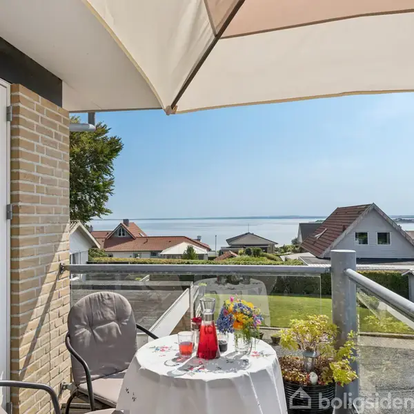 En terrasse med bord og stole er omgivet af blomster og skærmparasol. Bordet har en dug med blomster og en rød drink. Udsigt over huse og hav i baggrunden.