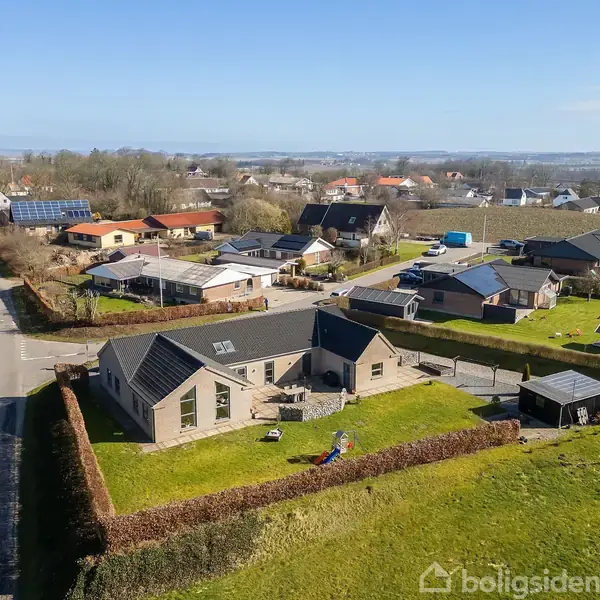 Hus med sort tag står stille inden for en hækindhegnet have. Omgivet af flere huse og veje i en landlig by. Blå himmel i baggrunden.