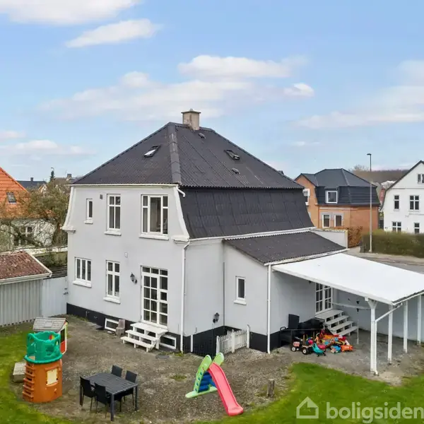 Et hvidt to-etagers hus med sort tag står i en have. En lille rutsjebane og legehus ses; en carport er ved siden af. Omkringliggende huse er synlige i baggrunden.