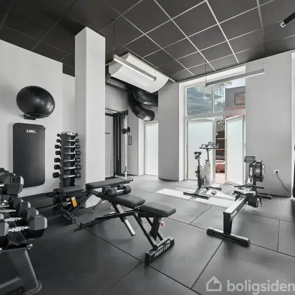 Et fitnesscenter fyldt med træningsudstyr som håndvægte, bænke, motionscykler og et løbebånd. Rummet har sorte gulve og lofter med lysindfald fra store vinduer.