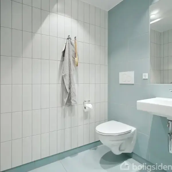 Toilet hænger på en flisevæg, ved siden af et vægmonteret sink. Et brusebad ses til venstre, og håndklæde hænger ved siden af. Miljøet er enkelt og moderne.