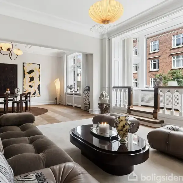 Stue med brun sofa placeret foran et sofabord med dekorative genstande, omgivet af store vinduer med udsigt til en murstensbygning. Spisebord ses i baggrunden med moderne kunst på væggen.