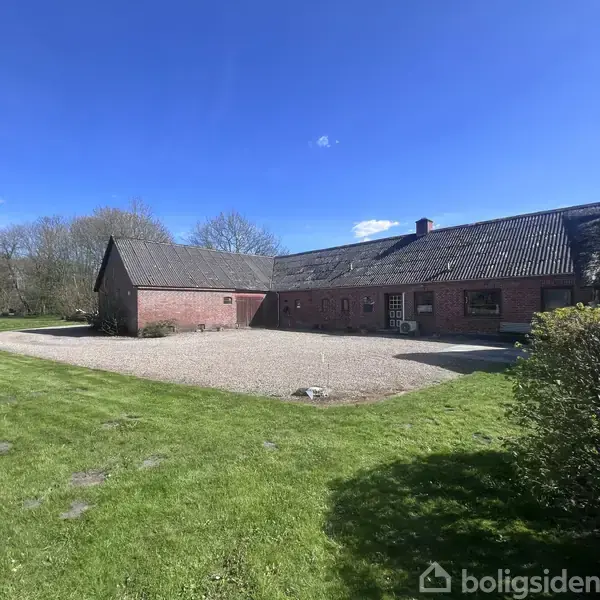 En rød murstensbygning står stille på en gårdsplads med grus, omgivet af grønne græsplæner og buske, under en klar blå himmel med spredte skyer.