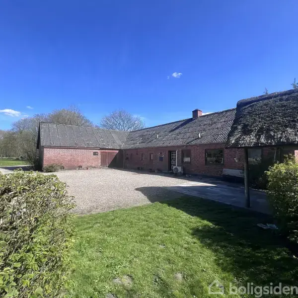 Rødt murstenshus med stråtag står tomt i en solrig gårdsplads omgivet af grønne buske og græsarealer under en klar blå himmel.