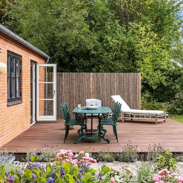 Terrassen består af et bord med stole og en liggestol på et trædæk. Den grænser op til et hus med røde mursten, omgivet af en frodig have med blomster.