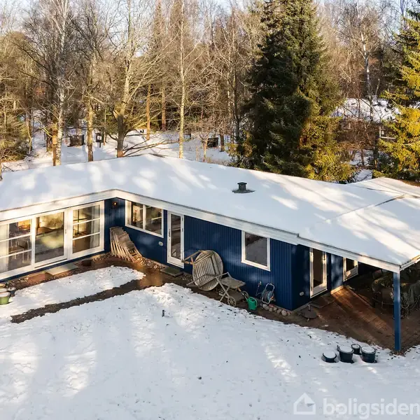 Et blåt hus med fladt tag er delvist dækket af sne. Det er beliggende i en skovklædt område med træer omkring, der giver en vinterlig atmosfære.
