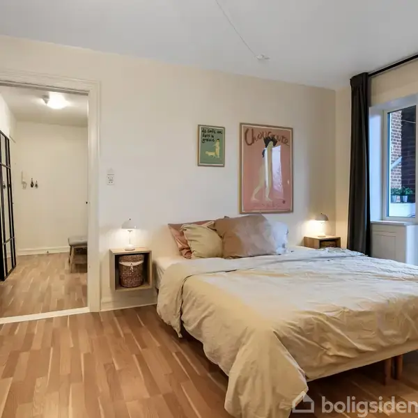 Soveværelse med en dobbeltseng dækket af beige sengetøj, omgivet af natborde. To kunstværker hænger på væggen. Rummet har trægulv og adgang til en balkon med glasdør.