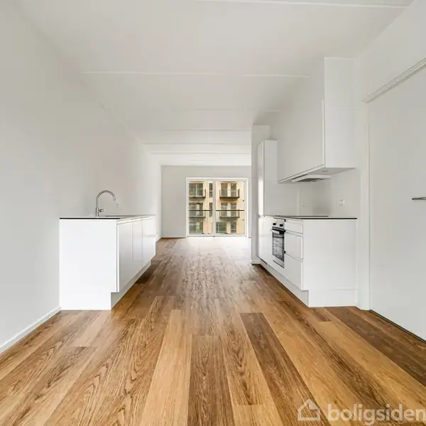 Et moderne køkken med hvide skabe og en ovn er forbundet til en stue, der har trægulve og udsigt til en balkon gennem store vinduer i baggrunden.