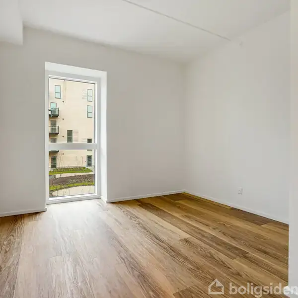Et lyst, tomt rum med træparketgulv og en enkelt glasdør, der åbner ud til en udsigt over en moderne bygning. Lokalet har hvide vægge.