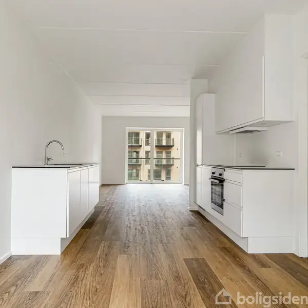 Et lyst, moderne køkken med hvide skabe og indbygget ovn eksisterer i et rum med trægulv. Køkkenet har udsigt til en balkon med glasdøre og udsigt til en bygning.