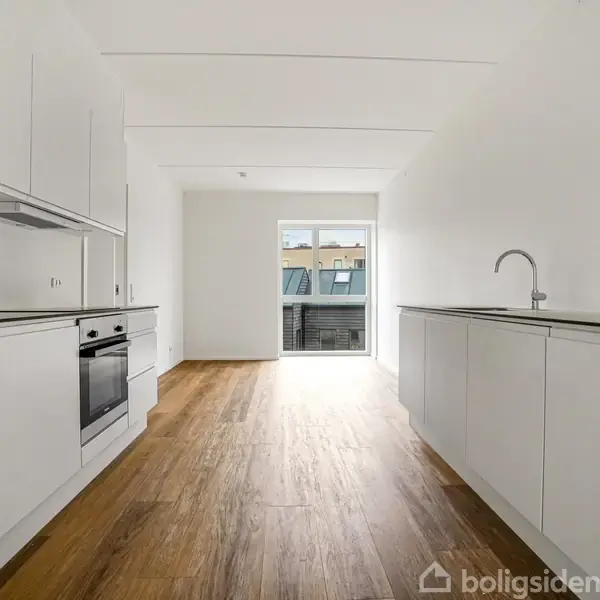 Et køkken med hvide skabe og en indbygget ovn ligger på begge sider af et langt rum med trægulv. I baggrunden er der en glasdør, som fører til en balkon.
