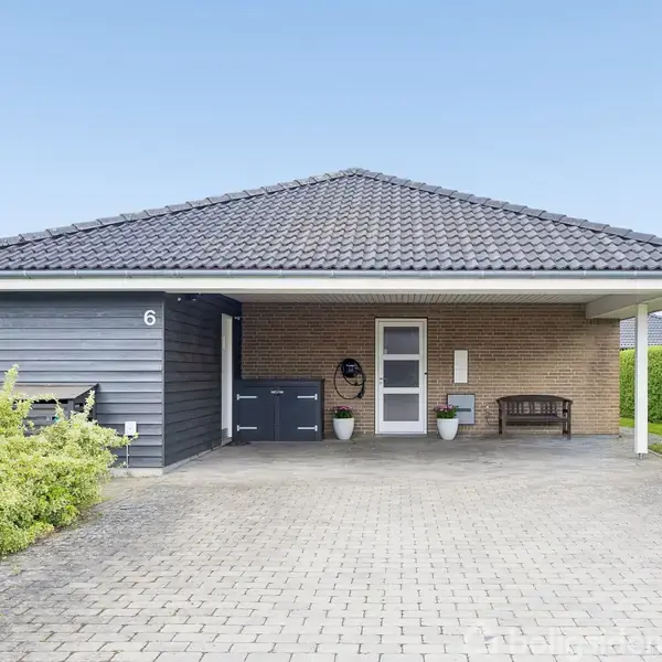 Hus står stille på en flisebelagt indkørsel med carport. Murstensvægge og sort tegltag. Grønne buske omgiver stedet. Skilt med teksten "REAL Mæglerne Til Salg" og tallet "6" ses.
